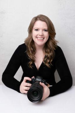 A lonfon portrait photographer with a camera in a black top against a white backdrop