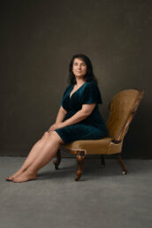A woman sitting on a gold chair in a green velvet dress sitting for a female portraits photography studio session