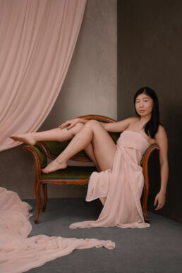An asian boudoir photograph with a woman seated on a couch in pink drapery