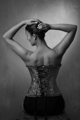 an indian woman raising her arms in a corset sitting on a vintage chair on a London boudoir photoshoot