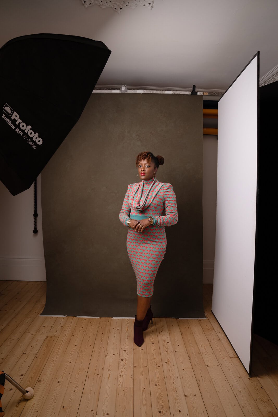 Portrait of a Black woman in a bold dress posing in a photography studio with green canvas, lights, and v-flat equipment visible.