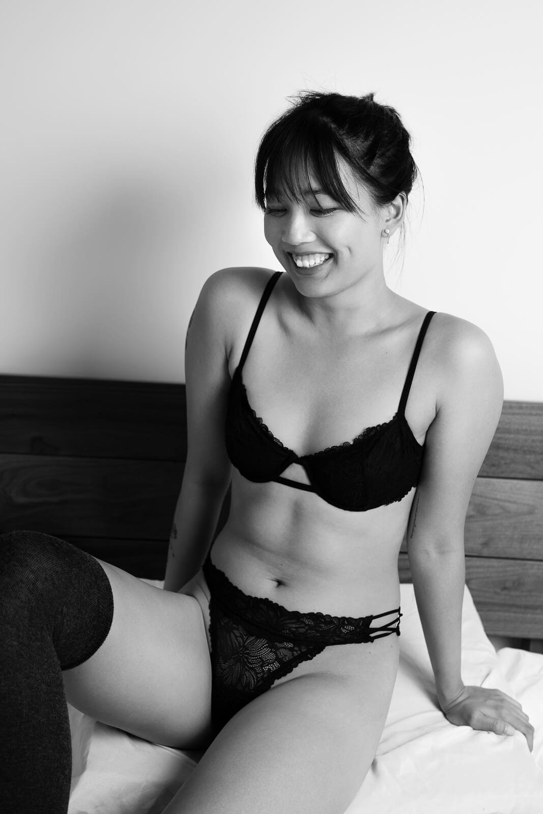 Asian woman laughing on a bed in lingerie, playful and relaxed moment captured as an intimate photograph.