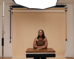 a black woman sitting at a posing desk surrounded by photography equipment in brown hues and taken by a london headshot photographer