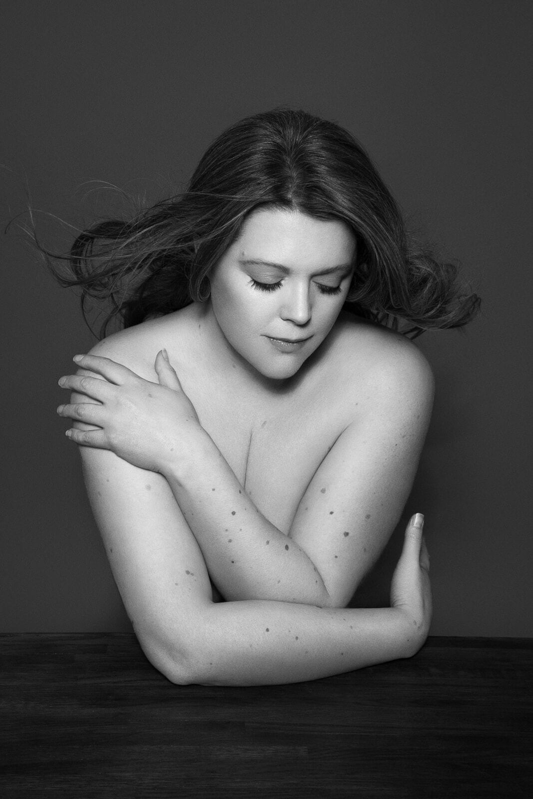 a female photographer in london holding her body at a table with hair blowing looking down in black and white