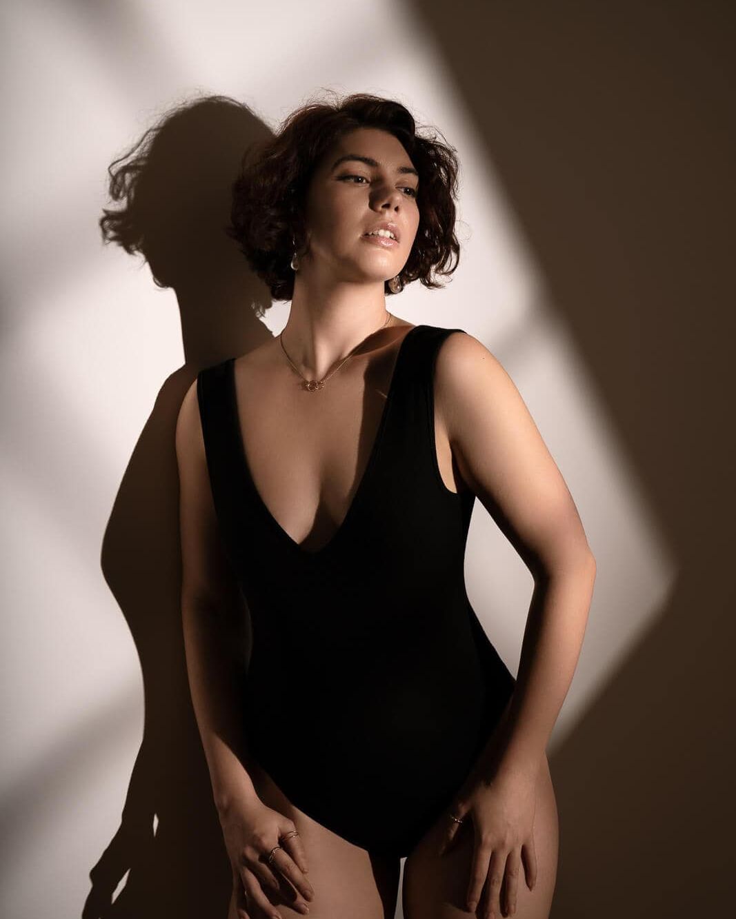 a woman in a black swim suit against a wall with window light on a sunlight boudoir photography experience
