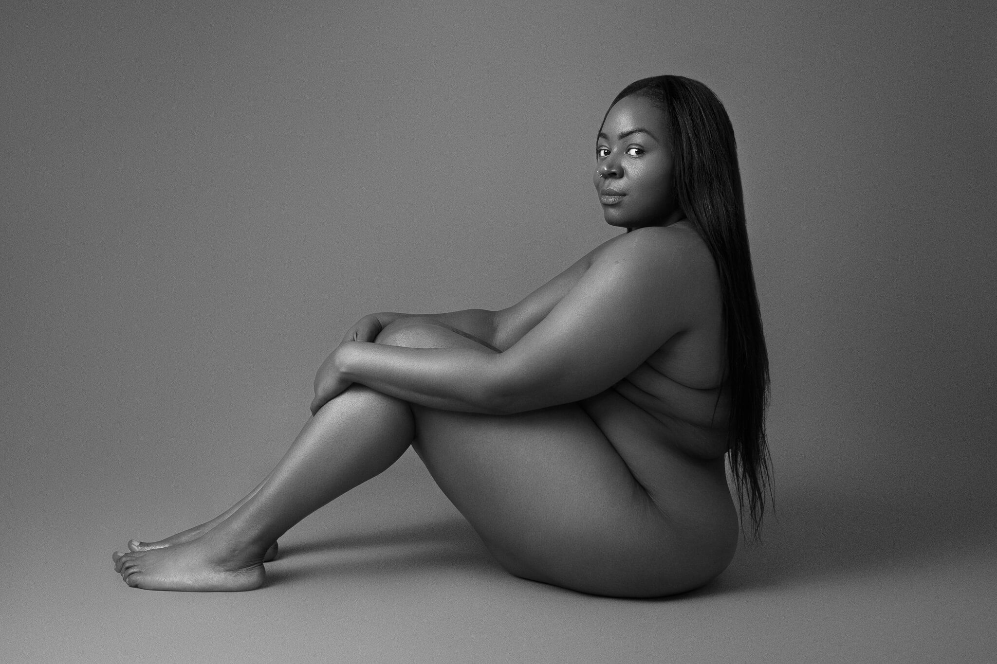 Black and white portrait of a Black woman seated and holding her knees, looking at the camera with calm defiance, representing the female gaze in boudoir photography.