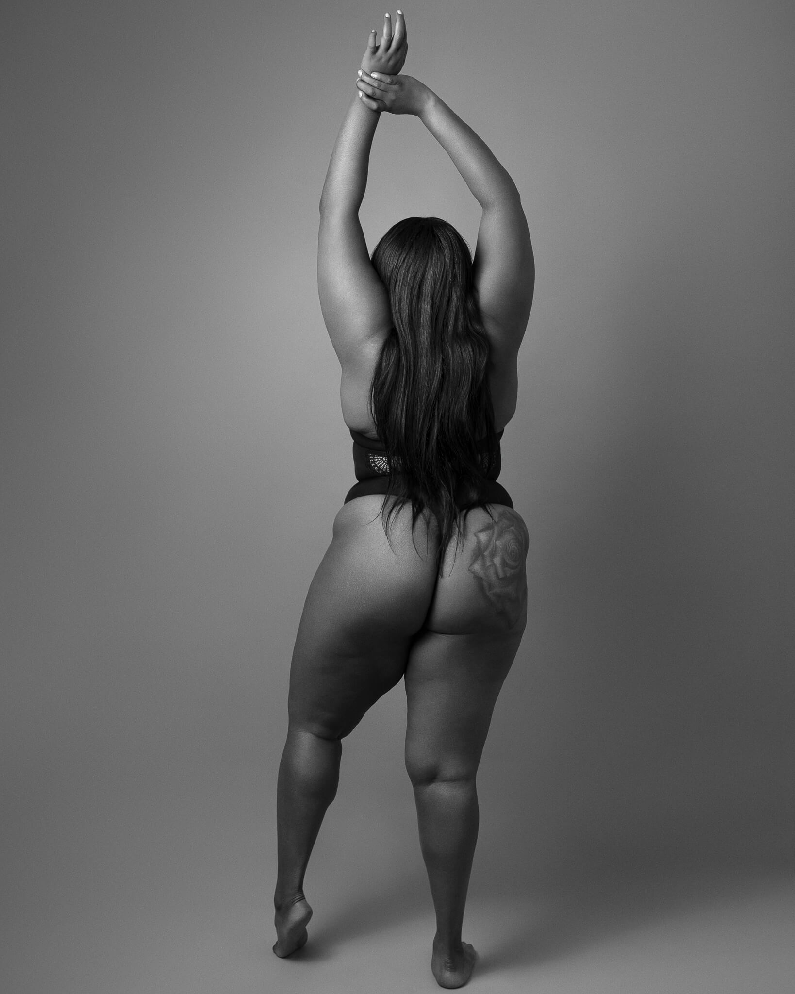 Black and white photograph of a Black woman seen from behind, arms raised and legs in motion, symbolising strength, freedom, and the female gaze in boudoir photography.