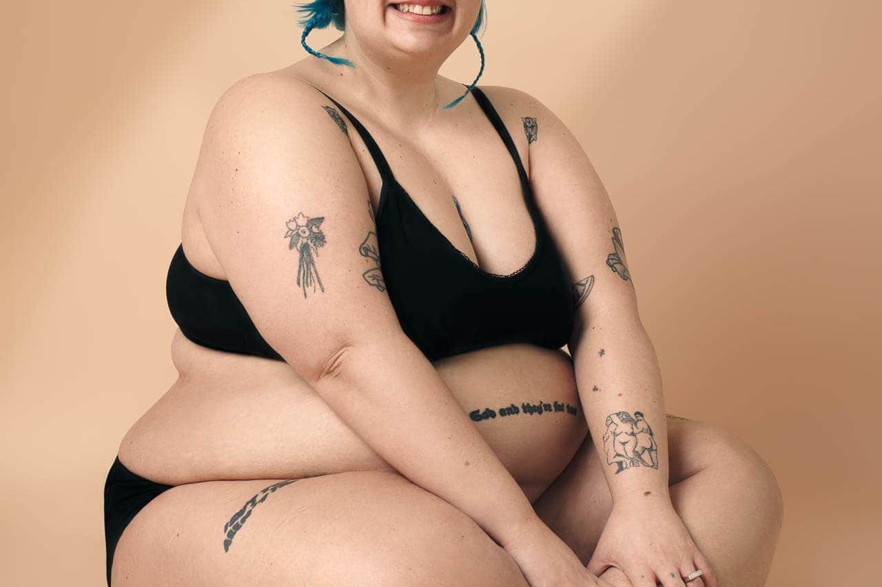Person with bright blue hair sitting cross-legged in black underwear against a warm beige backdrop during a private boudoir photoshoot in London.