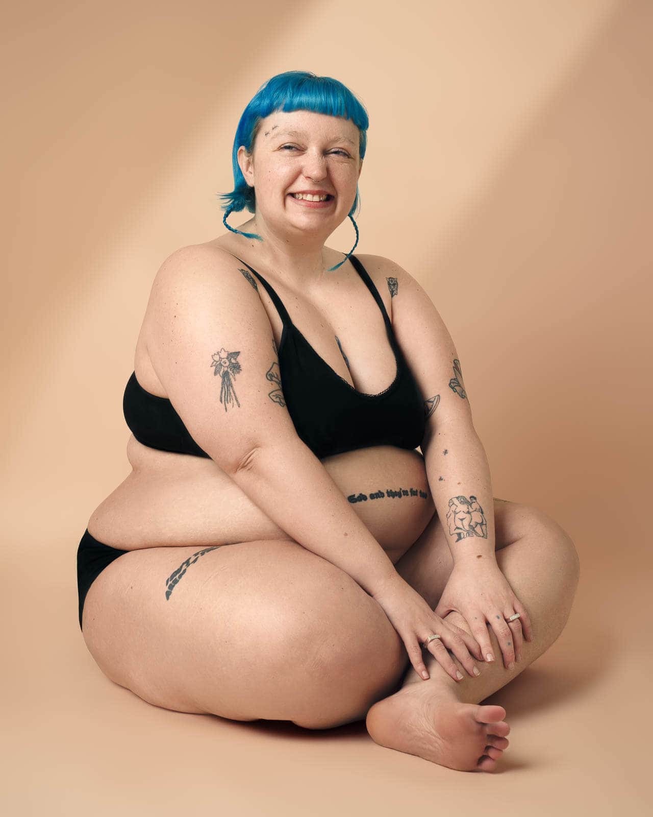 Person with bright blue hair sitting cross-legged in black underwear against a warm beige backdrop during a private boudoir photoshoot in London.