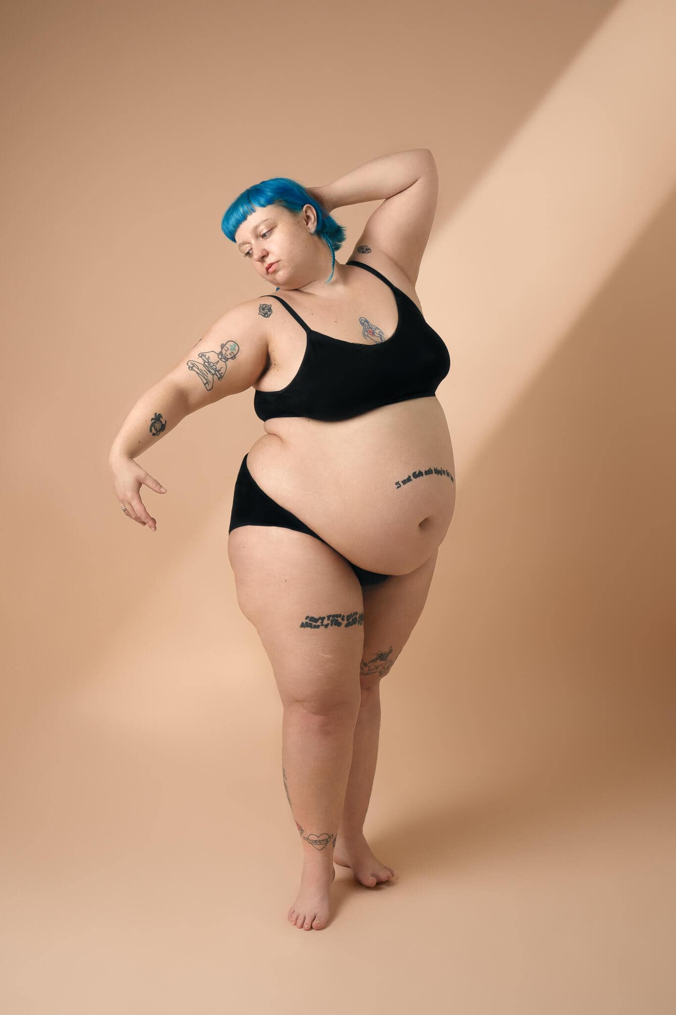 Person with bright blue hair posing in black underwear against a beige backdrop during a guided boudoir session in London.