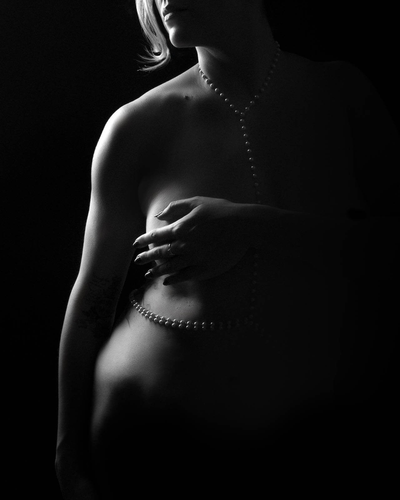 Black and white boudoir portrait of a partially lit figure wearing pearls, photographed in shadow with an intimate and private feel.