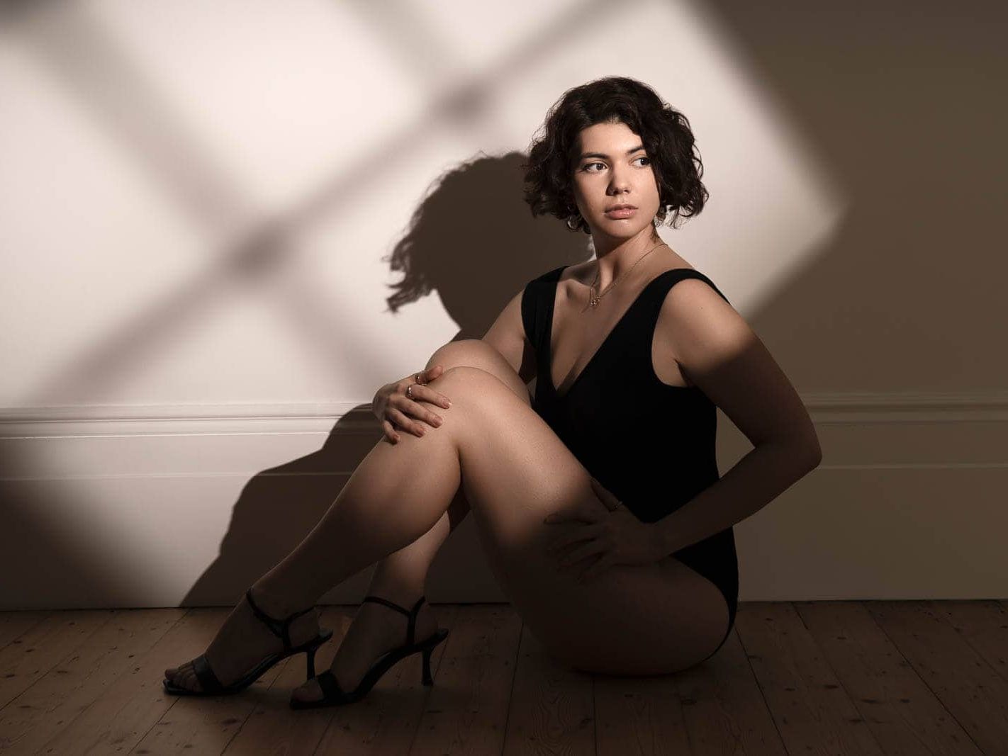 Person seated on a wooden floor in a black bodysuit and heels, photographed in warm sunlight during a boudoir session in London.