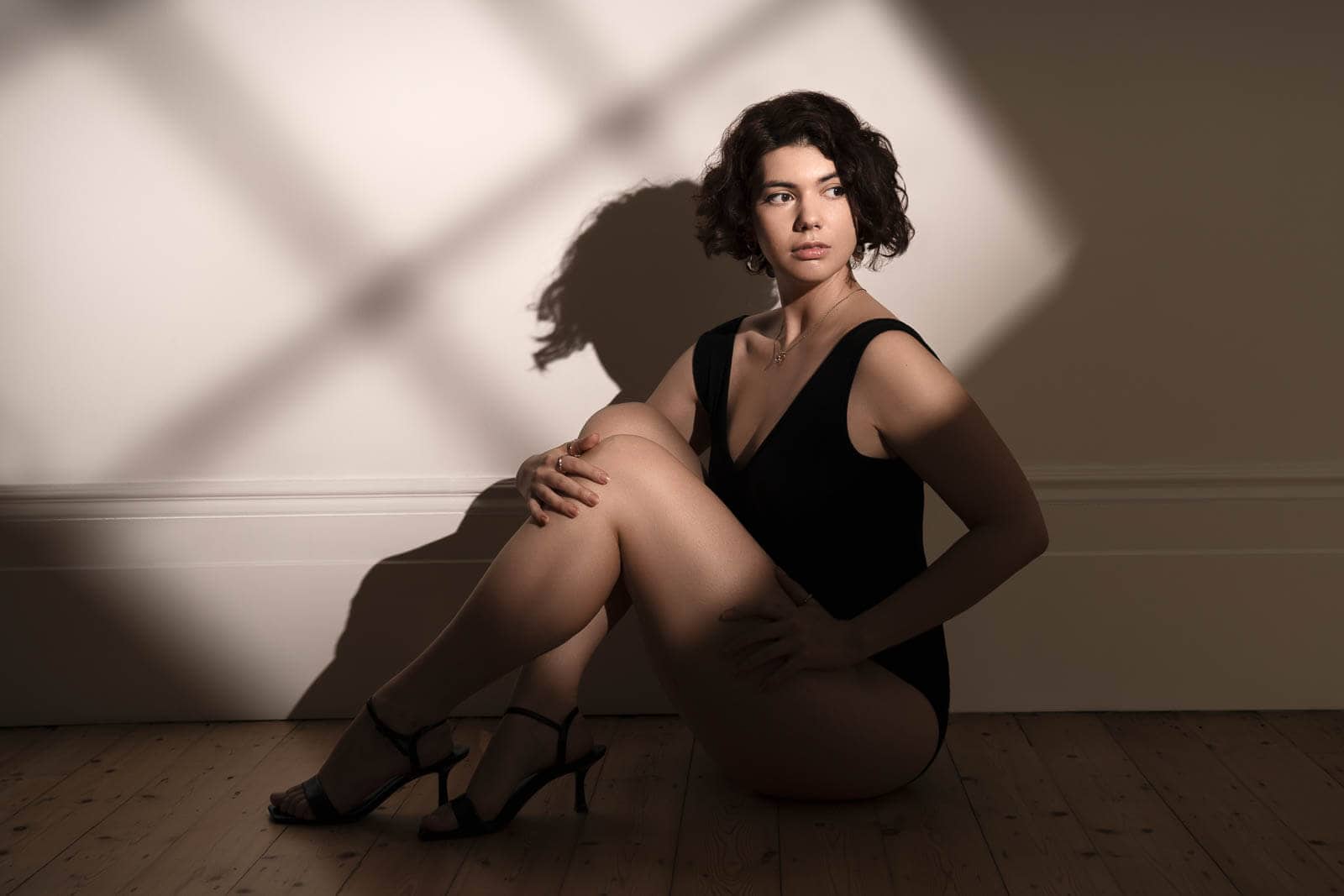 Person seated on a wooden floor in a black bodysuit and heels, photographed in warm sunlight during a boudoir session in London.