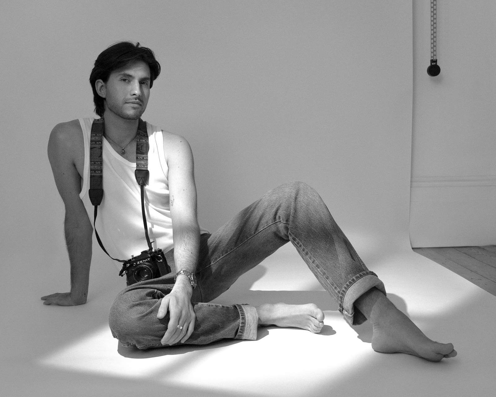 Black-and-white portrait of a man seated in sunlight with a vintage camera around his neck.